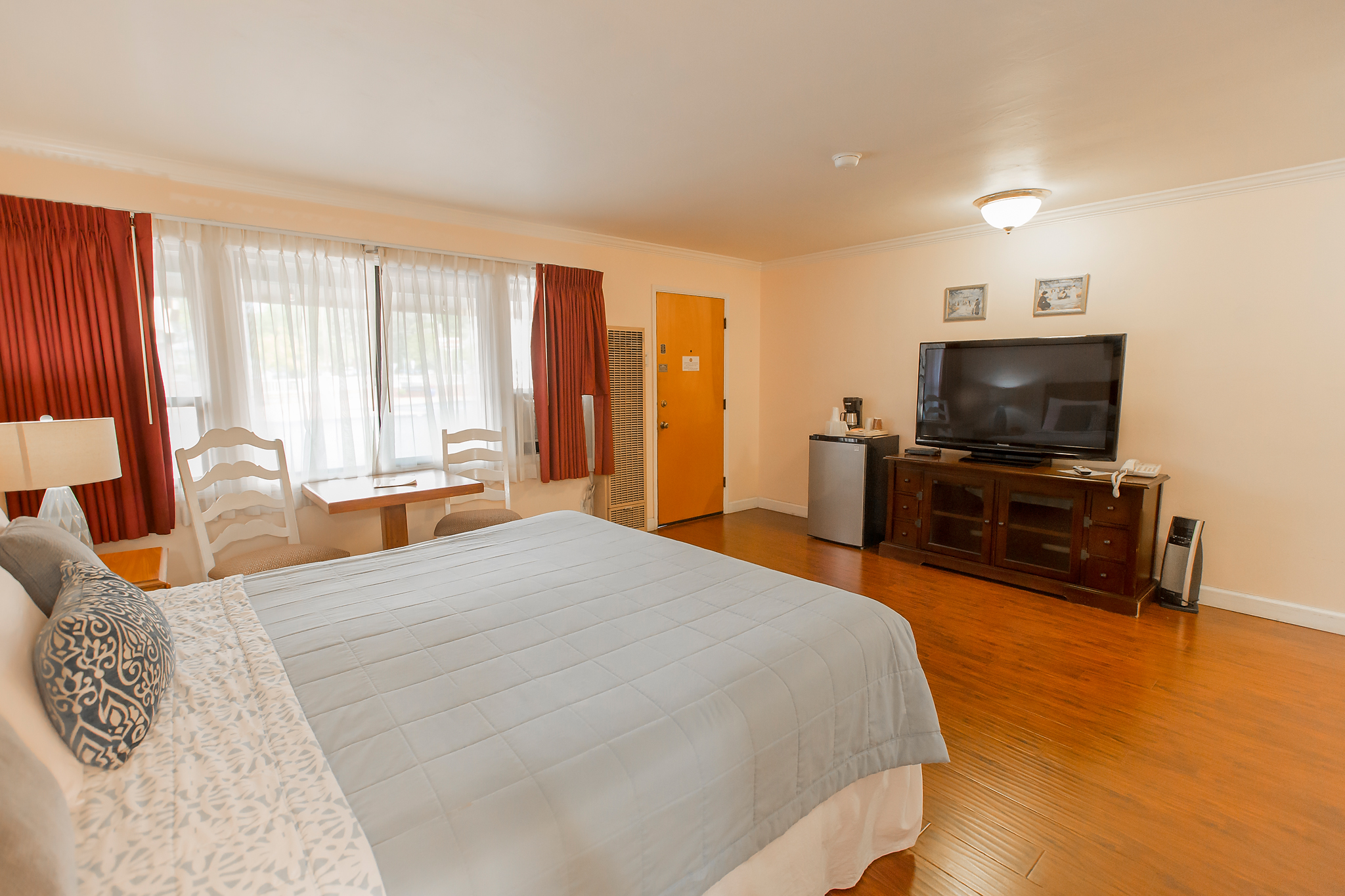 A wide view of a hotel room with a large bed, TV on a stand, table and chairs, and a window with red curtains.