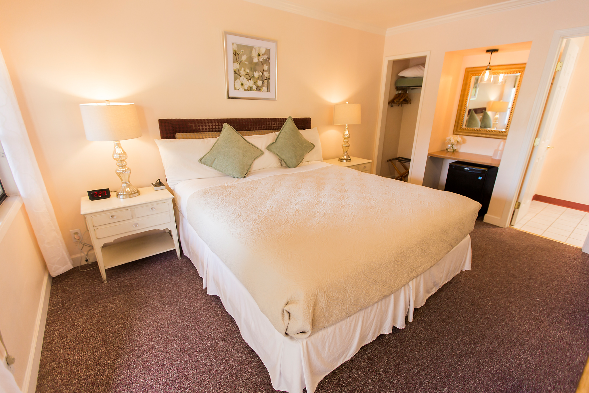 A wide-angle view of a hotel room with a queen bed, nightstand, lamp, closet, and mini fridge.