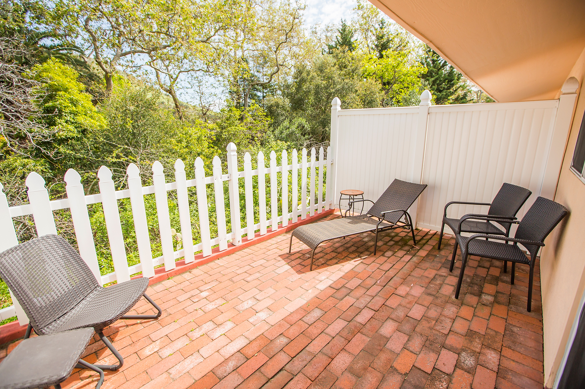 A brick patio with outdoor furniture and a white picket fence, surrounded by lush greenery.