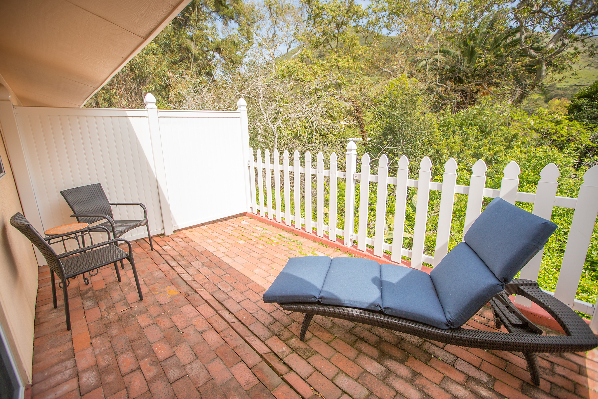 A brick patio with a lounge chair, small table, and white picket fence.